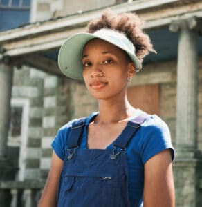 Black woman wearing a blue shirt, denim overalls and a white visor stands in front of a stone building with Grecian-style columns