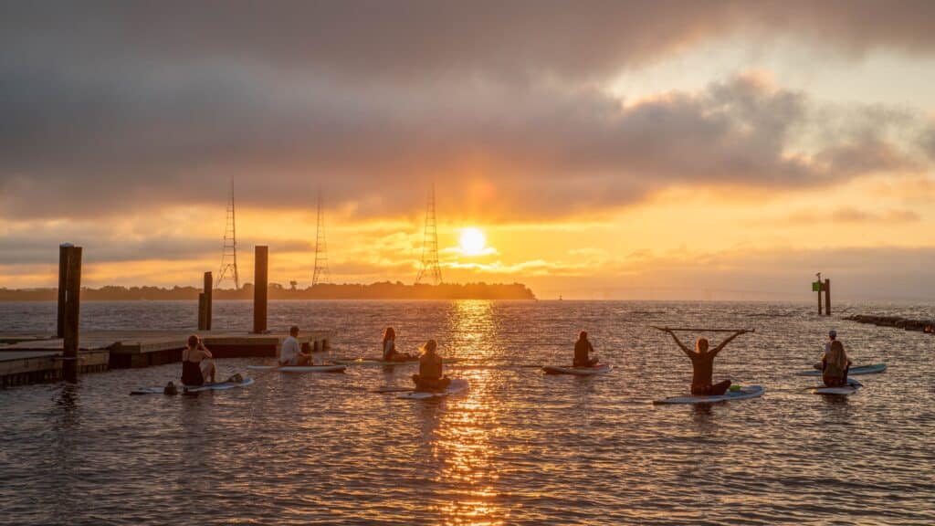 Group of seven people sitting on stand up paddleboards on the water in front of a sunrise