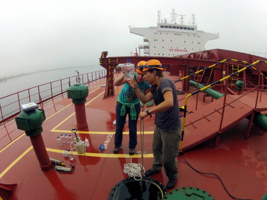 Two scientists in hard hats stand on the red deck of a giant commercial ship, looking at a water sample in a clear bottle.