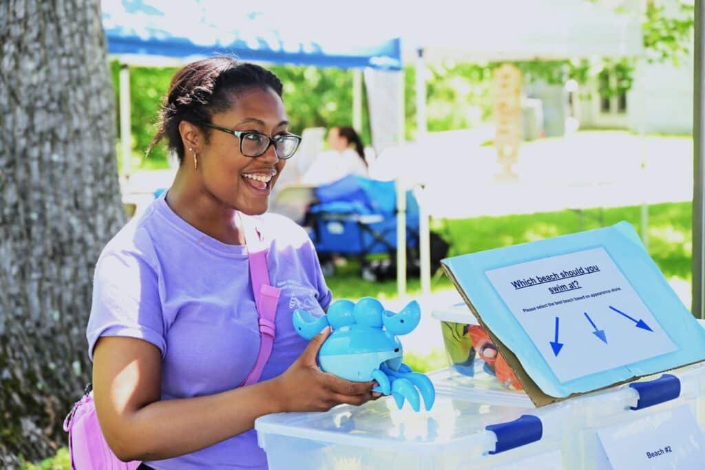 An individual in a light purple T-shirt stands behind an outdoor display, holding a plastic blue crab and smiling widely. A blue sign on the display table reads, Which beach should you swim at