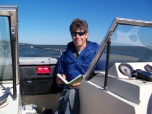 Whitman Milller, wearing sunglasses and a blue windbreaker sits on a boat, holding a book and smiling at the camera.