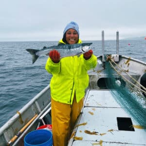 Imani Black, wearing a bright yellow windbreaker and red gloves, stands on a fishing boat holding a large silver fish in both hands and smiling at the camera